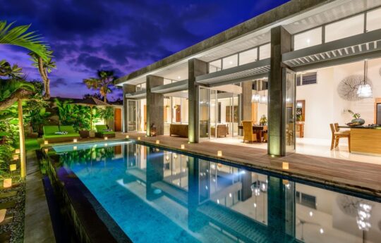 Villa Bamboo swimming pool and sunbeds with view of interiors at night with lights on