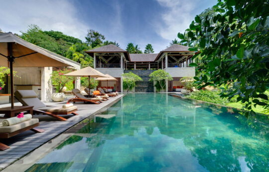 Large swimming pool with garden and villa exteriors with day beds by the pool on sunny day