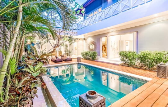 Swimming pool, deck area with plants in foreground and villa exterior at night with lights on