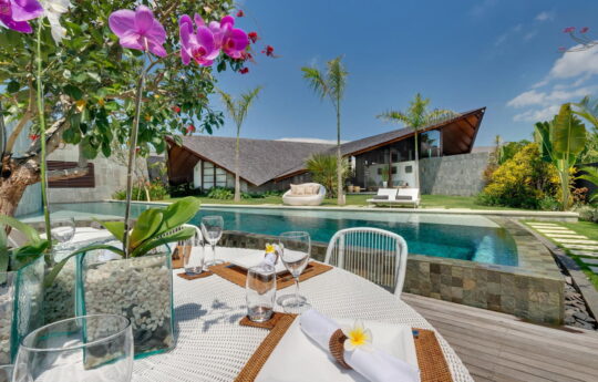 Swimming pool on sunny day with villa in background and breakfast table in foreground